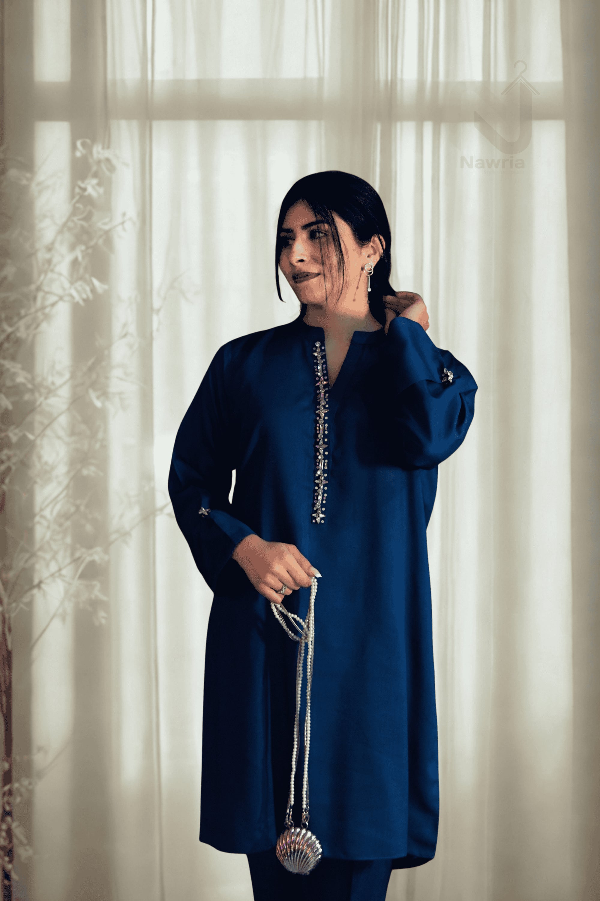 Woman wearing a blue traditional outfit with a decorative pendant, standing in front of light curtains.